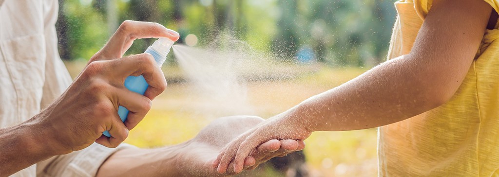 Persona aplicando repelente de insectos en spray sobre el brazo de un niño, al aire libre en un día soleado, rodeados de vegetación.