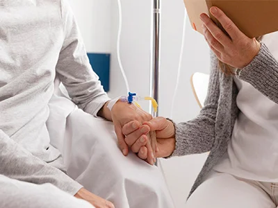 Paciente oncológico recibiendo tratamiento intravenoso mientras es acompañado por una persona que le sostiene la mano. Simboliza el apoyo emocional y la importancia del acompañamiento en el proceso de lucha contra el cáncer.