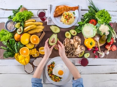 Vista cenital de mesa con alimentos saludables como frutas, verduras, aguacate, salmón y huevo, y una persona partiendo una palta/aguacata, destacando una dieta equilibrada.