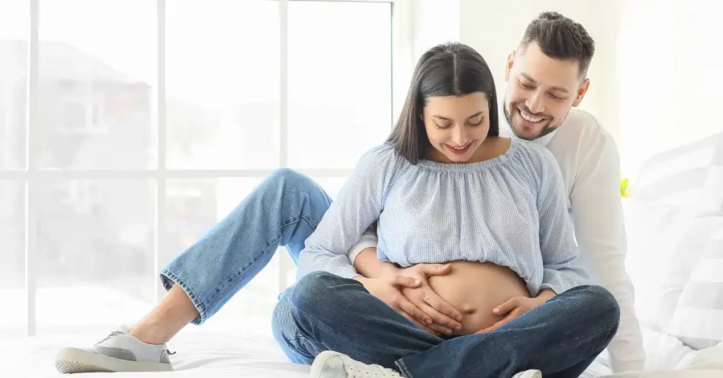 Pareja sentada enel suelo sonriendo, mientras el hombre abraza a la mujer embarazada, haciendo referencia a fertilidad humana