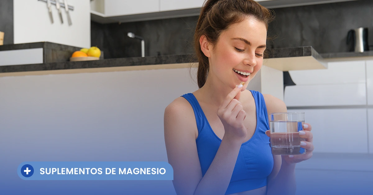 Mujer joven sonriendo en su cocina mientras se prepara para tomar una cápsula con un vaso de agua, ilustrando el uso de los distintos tipos de magnesio en suplementos.