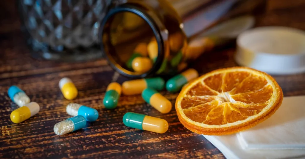 frasco de pastillas volcado con cápsulas de colores dispersas sobre una mesa de madera, junto a una rodaja de naranja seca, representando las Vitaminas para diabéticos y los suplementos alimenticios.