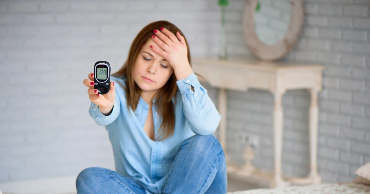 Una mujer joven con expresión angustiada y la mano en la frente, sosteniendo un medidor de glucosa digital preocupada por los síntomas de la diabetes