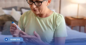 Una mujer mayor con gafas, sentada en una habitación, sostiene un dispositivo de punción cerca de su dedo para el control glucémico. En la esquina inferior izquierda, un banner azul muestra el texto 'DIABETES', contextualizando el manejo de esta patología metabólica, la cual es frecuentemente abordada con tratamientos del tipo GLP-1
