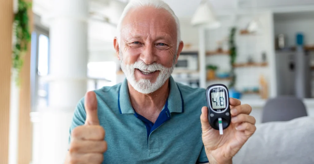 Hombre mayor sonriendo en su hogar, sosteniendo un glucómetro y levantando el pulgar en señal de mantener una glucosa normal en adultos.